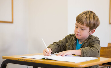 Boy student wearing olive jacket writing in open workbook at school desk, holding white pen