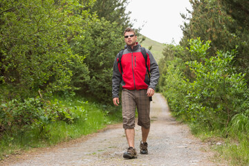 Fototapeta premium Man walking gravel trail through woods, wearing red-gray jacket, hiking shoes, backpack, sunglasses