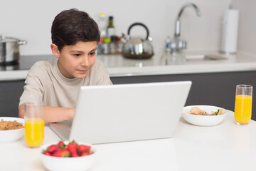 School-age boy sitting at counter using silver laptop with strawberries and juice in home kitchen
