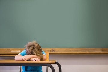 Boy child resting head on arms at desk in classroom with green chalkboard, wooden chalk tray