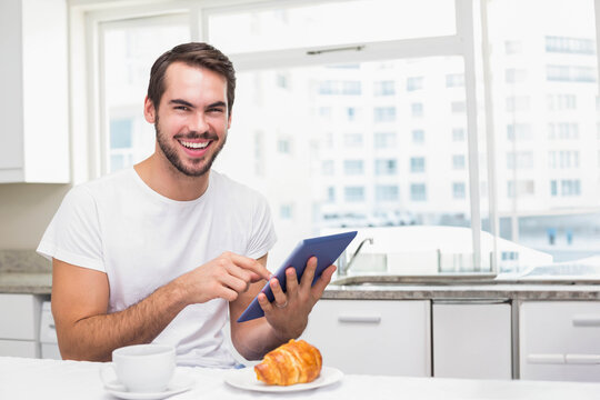 Man sitting at kitchen table wearing white T-shirt, holding blue tablet, smiling near coffee cup