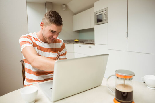 Male sitting at kitchen table working on silver laptop in orange striped t-shirt with French press