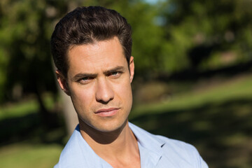 Mid-30s male standing posed in sunny park wearing light blue collared shirt with blurred background