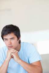 Man sitting leaning clasping hands under chin in living room near sofa back wearing blue T-shirt