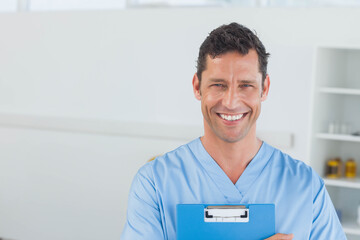 Older man smiling holding clipboard wearing blue scrubs at clinic, amber meds on shelf, copy space