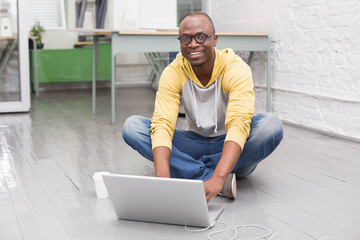 African American man sitting in studio working on silver laptop, yellow hoodie, glasses, coffee cup