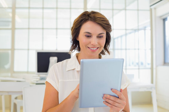 Mature woman holding tablet while standing in white blouse at open-plan office near grid windows