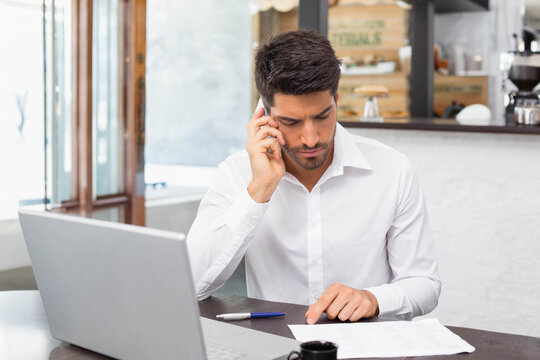 Late 20s man in white shirt sitting at cafe reading documents with laptop, phone, blue-pen, coffee