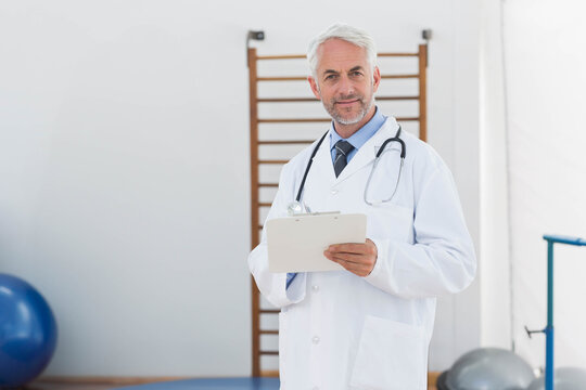 Male therapist in white lab coat with stethoscope holding clipboard in rehab near exercise balls