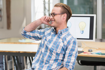 Man phoning, smiling, sitting at wood desk in open office with monitor lightbulbs and swatches