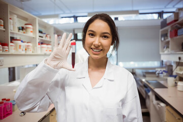 Asian woman wearing white lab coat and glove holding capped blood collection tube at lab bench