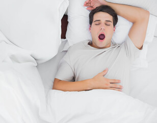 Man lying on bed yawning, hand on chest, wearing gray T-shirt amid white pillows and duvet