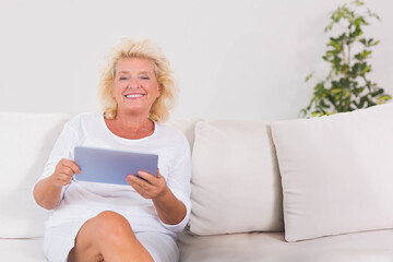 Senior woman sitting on sofa in living room holding tablet wearing white outfit, copy space
