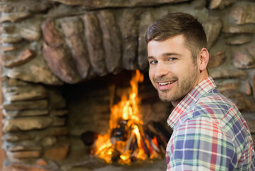 Naklejka premium Adult bearded man smiling, sitting by stone fireplace burning logs, wearing plaid shirt, copy space