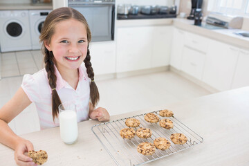Schoolgirl in pink shirt smiling, holding cookie at kitchen with milk, cooling rack, copy space