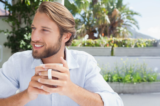 Man sitting on sunny terrace holding white ceramic mug at table, wearing shirt, planters with palms