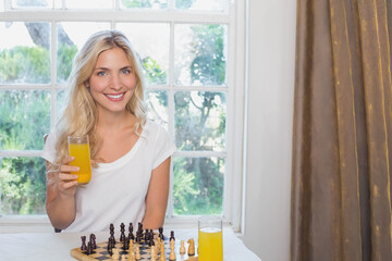 Woman sitting at home table holding orange juice smiling at camera chessboard by window curtain