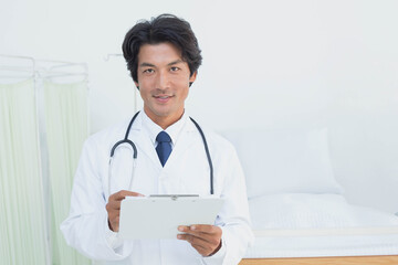 Asian mid-adult male doctor in lab coat with stethoscope writing on clipboard with pen in clinic
