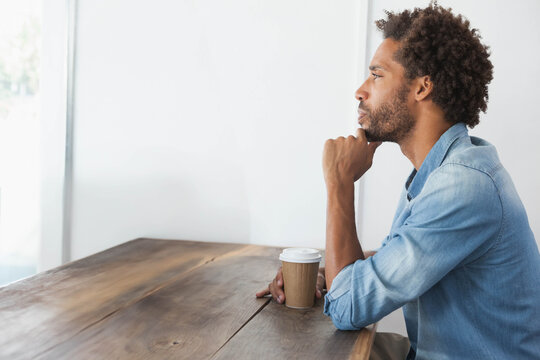 Mid adult African American man sitting at wood table holding coffee cup, gazing right, copy space - Powered by Adobe
