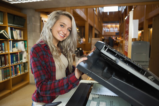 Adult woman in plaid shirt lifting copier lid and placing spiral notebook at library near shelves