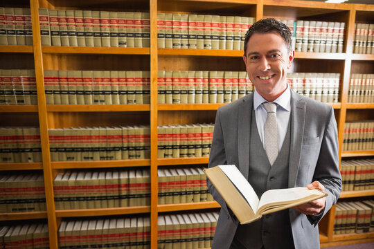 Man in suit standing by wooden bookshelves in law library holding open legal volume, copy space - Powered by Adobe