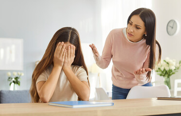 Upset mother scolding teenage daughter sitting at table during family conflict. Sad girl covering face with hands, showing distress, while parent expressing frustration during emotional conversation.