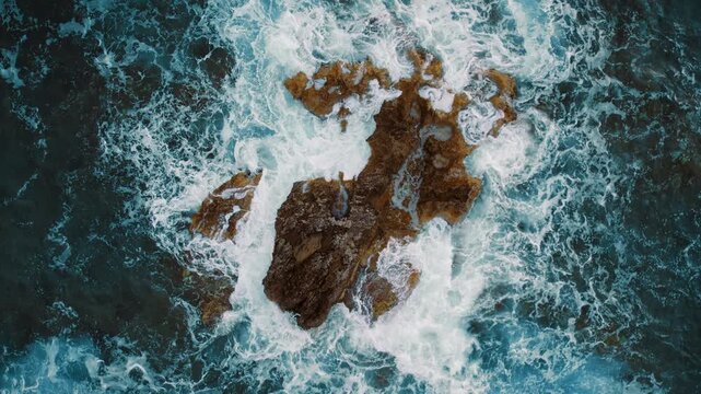 A rocky crest pierces foamy blue as sea waves beat in tidal chaos below