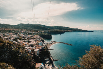 Wide aerial view of the coastal town, waterfront structures, small beach, and turquoise Mediterranean Sea under a partly cloudy sky.