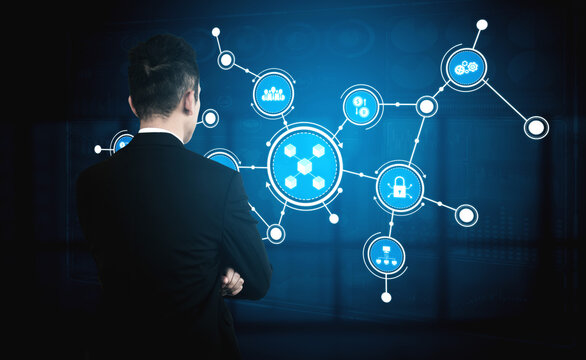 A business professional stands in a dark office, analyzing a futuristic digital network on an interactive screen, featuring blue graphics and various technology icons. Tessel - Powered by Adobe