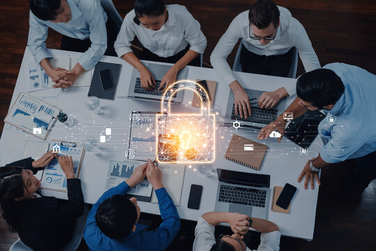 A diverse team meets around a table with laptops and documents, discussing data security highlighted by a digital lock graphic, emphasizing the importance of information protection. Trope - Powered by Adobe