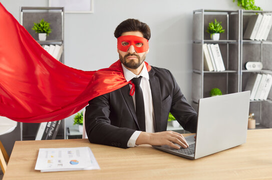 Business man in formal suit and superhero face mask and cloak working in office with laptop. Portrait of a funny confident successful male company employee sitting at the desk on workplace.
