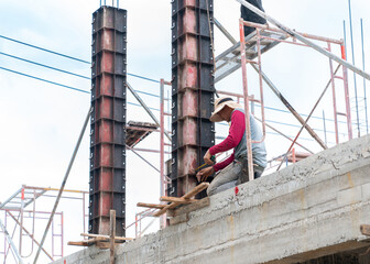 Naklejka premium A construction worker, wearing a long-sleeved shirt and a wide-brimmed sun hat, crouches on a newly poured concrete beam or slab.