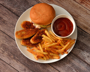 A fast food lunch: a hamburger, chicken strips, and French fries with tomato sauce on a round plate on a wooden table.