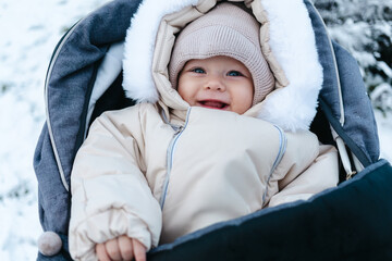 Happy smiling baby boy 11 months old wearing beige overalls and warm knitted hat, walking outside in winter is sitting in a grey baby stroller and looking on the snow. Park in the background