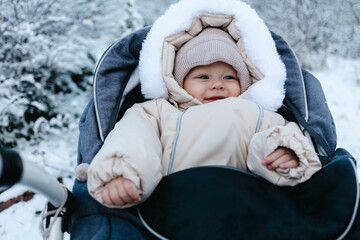 Happy smiling baby boy 11 months old wearing beige overalls and warm knitted hat, walking outside in winter is sitting in a grey baby stroller and looking on the snow. Park in the background