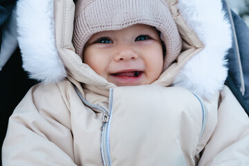 Happy smiling baby boy 11 months old wearing beige overalls and warm knitted hat, walking outside in winter is sitting in a grey baby stroller and looking on the snow. Park in the background