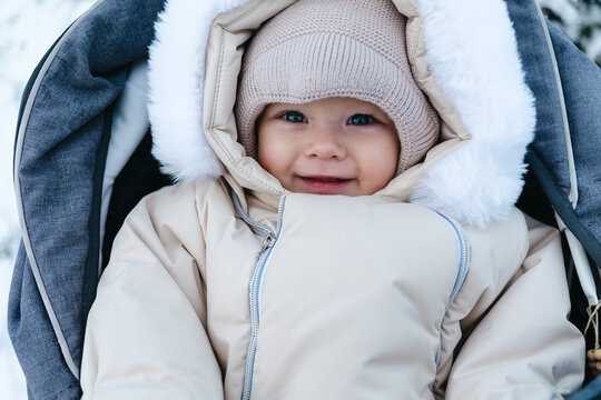 Happy smiling baby boy 11 months old wearing beige overalls and warm knitted hat, walking outside in winter is sitting in a grey baby stroller and looking on the snow. Park in the background