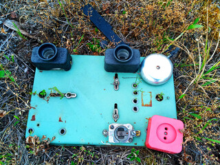 A rusty, turquoise electrical panel with a bell, socket, and lamp holders lies abandoned on the ground