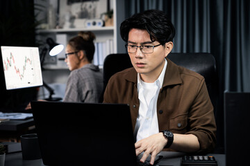 Serious face of thinking businessman focusing laptop to report data analysis social media online project with coworker analyzing stock at back side at over late night time at modern office. Infobahn.