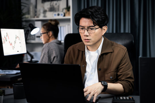 Serious face of thinking businessman focusing laptop to report data analysis social media online project with coworker analyzing stock at back side at over late night time at modern office. Infobahn.