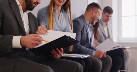 Group of corporate workers sitting in row and discussing something. Job applicants came on interview, sitting in line, holding their resumes, posing in business environment, talking with each other - Powered by Adobe