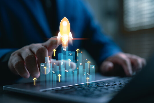 Startup business growth concept. Businessman pointing at rising bar graph with rocket icon, percentage sign and upward arrows, symbolizing success, investment return, and financial innovation.