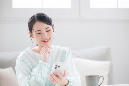 スマホ持つ女性が笑顔で幸せ感じる室内のかわいい日本人　問い合わせなどで考える　よくある質問のイメージ