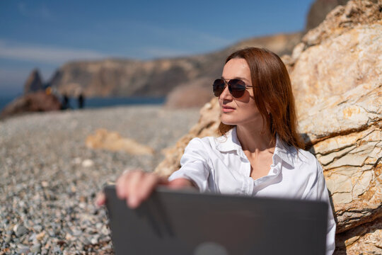 Woman remote work beach freelancer uses laptop near seaside cliffs under clear sky