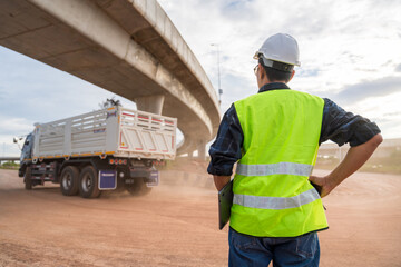 An engineer in safety vest and hard hat supervising dump truck operations on a dusty, unfinished highway construction site under an overpass.