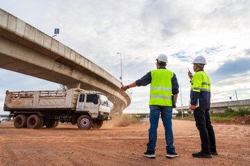 Two construction engineers, wearing safety gear, direct a dusty dump truck leaving the worksite under a large highway overpass.