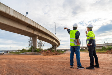 Two construction engineers stand on a muddy worksite, pointing and communicating via radio under a massive curved highway overpass.