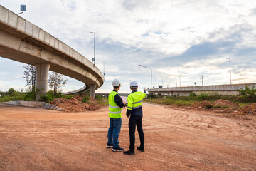 Two construction engineers stand on a vast dirt road, inspecting the site under a sweeping curved concrete highway overpass.