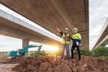 Two construction engineers stand on a dirt mound, communicating via radio and inspecting an excavator under a large overpass at sunset.
