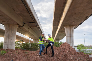 Two construction engineers stand on a high dirt pile, reviewing plans on a laptop under dual towering highway overpasses at sunset.